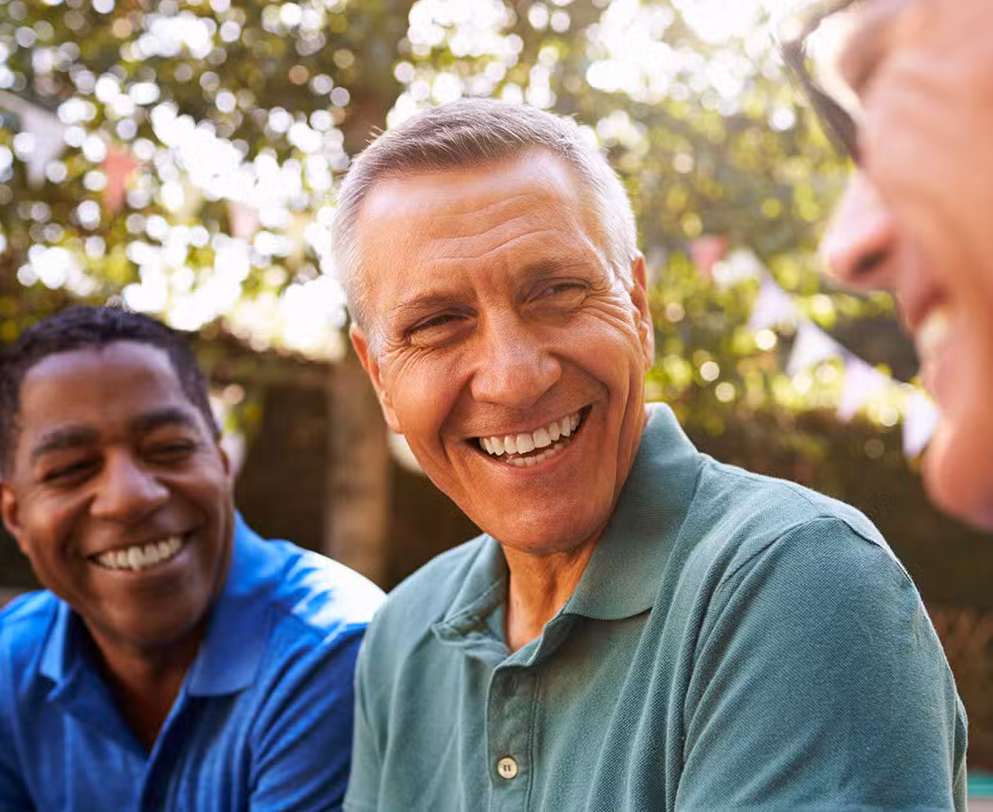 Three people sit outdoors, smiling and laughing together. The focus is on a middle-aged man in a green shirt, with two others‚Äîone in blue and one in glasses‚Äîenjoying the moment in a sunlit, leafy setting.