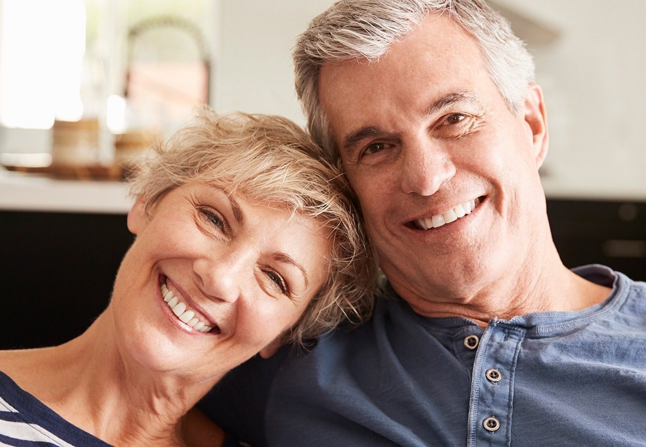 A smiling older couple with gray hair sits closely together indoors, looking at the camera. The woman leans her head on the man‚Äôs shoulder. They both wear casual shirts and appear happy and relaxed.